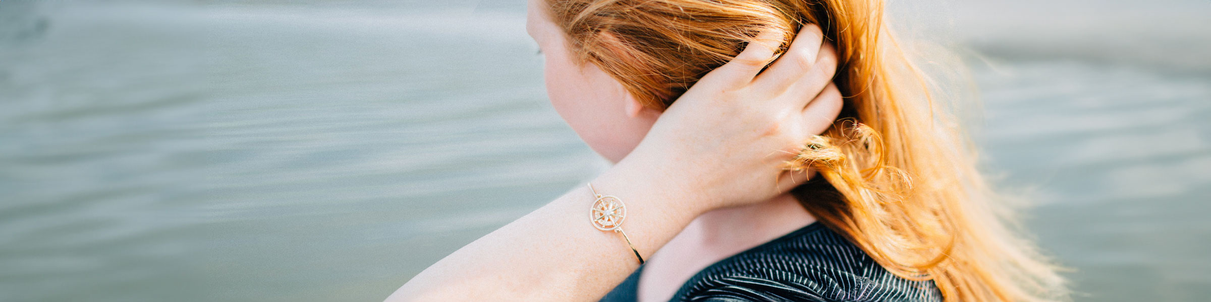 Woman with red hair looking out on water with gold compass rose bracelet